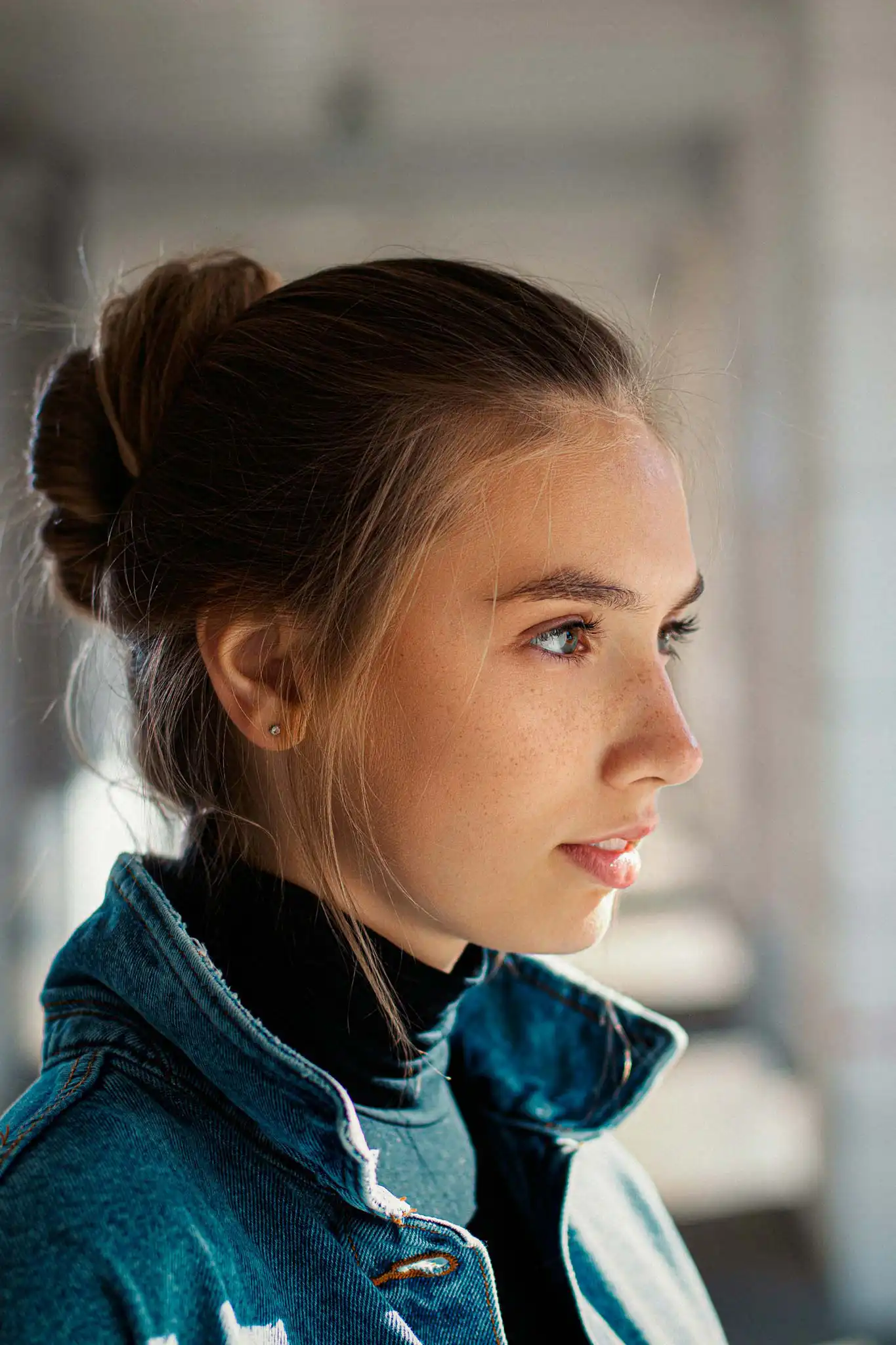 Close-up profile portrait of a young woman with her hair in a bun, wearing a denim jacket